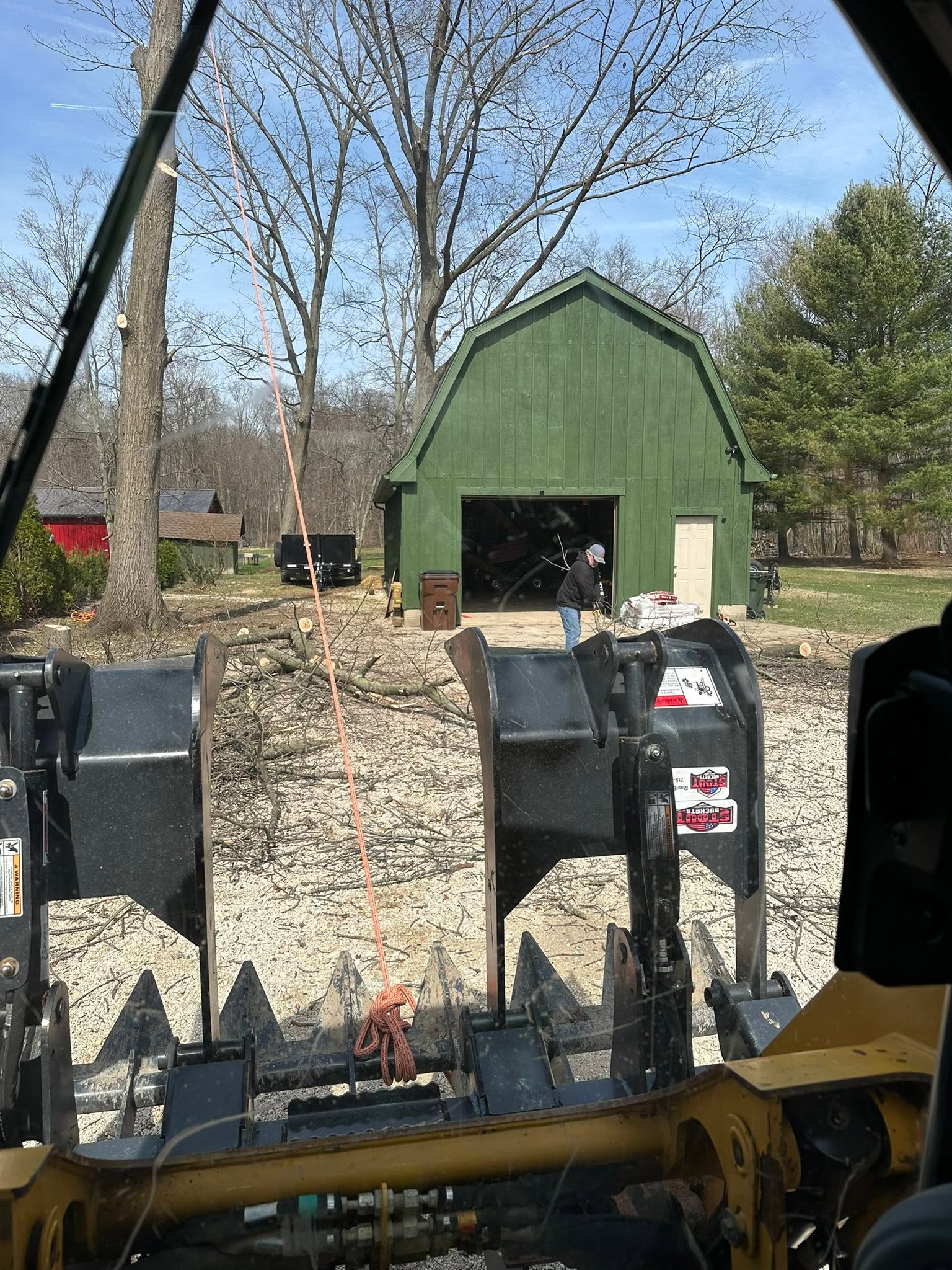 Skid steer clearing branches after tree removal near a green barn